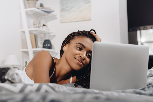 Mulher negra deitada de bruços na cama mexendo no computador. 